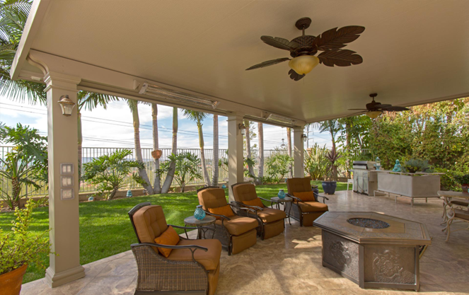 A patio with several outdoor reclining chairs and side tables around a stone fire pit with a patio cover overhead that is supported by thick columns and has two decorative ceiling fans suspended from it.